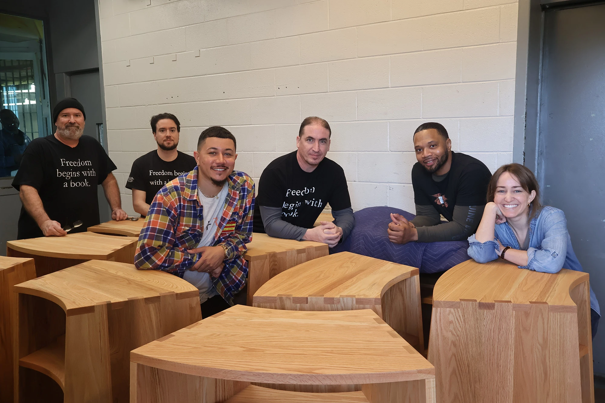 6 members of the Freedom Reads team pose for a photo around 9 wooden bookcases in a loading dock at Garden State Correctional Facility.