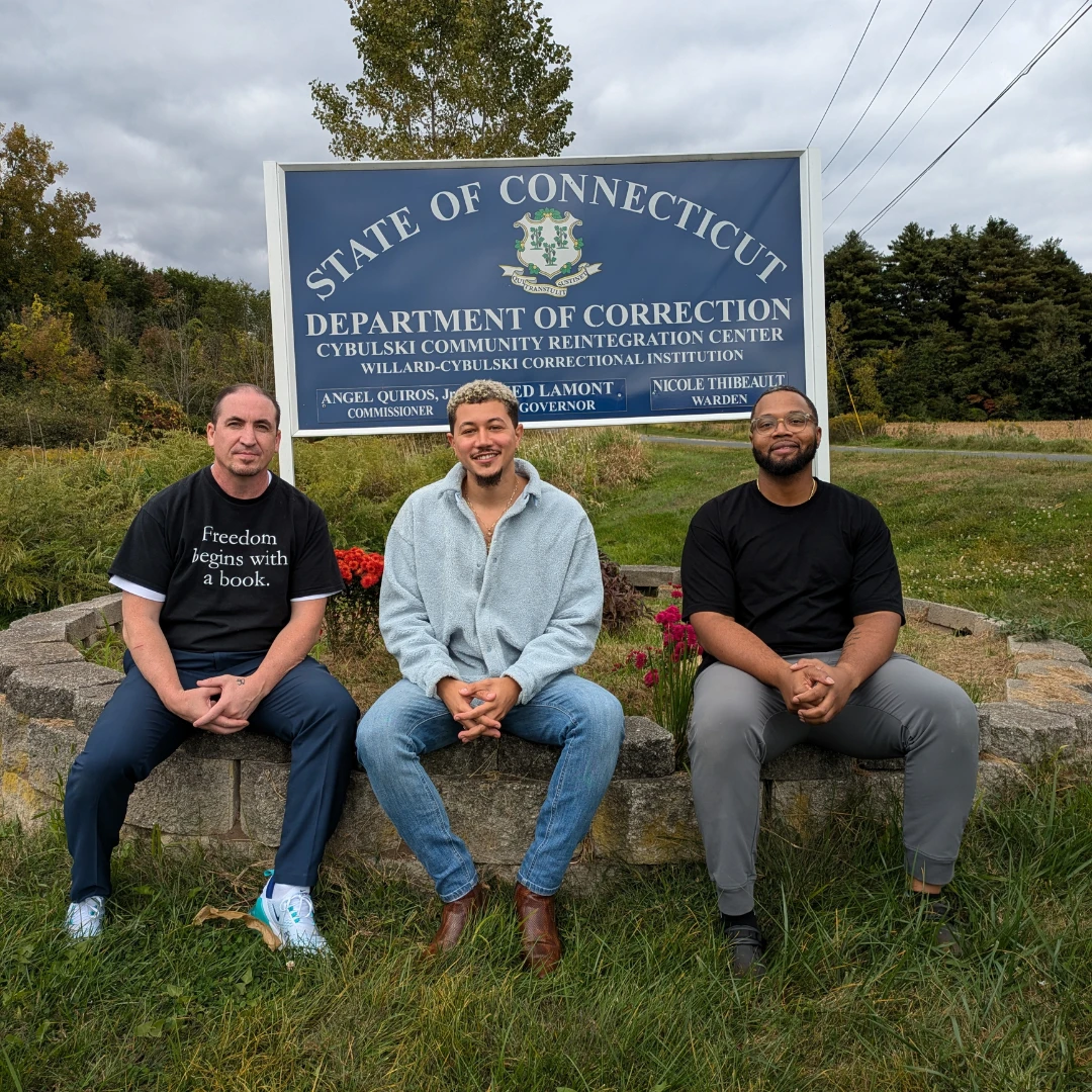 Freedom Reads Communications Manager Steven Parkhurst (left), Freedom Reads Library Coordinator David Perez Jr (center), and Freedom Reads Library Production Assistant Michael Byrd (right) sit outside of Cybulski Community Reintegration Center sign.