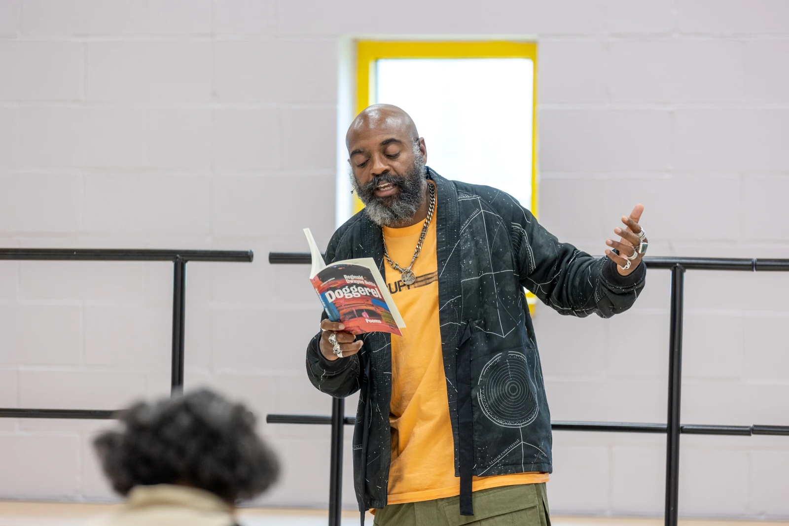 Dwayne wearing a yellow shirt and black jacket standing at the front of a room giving a poetry reading from his book doggerel