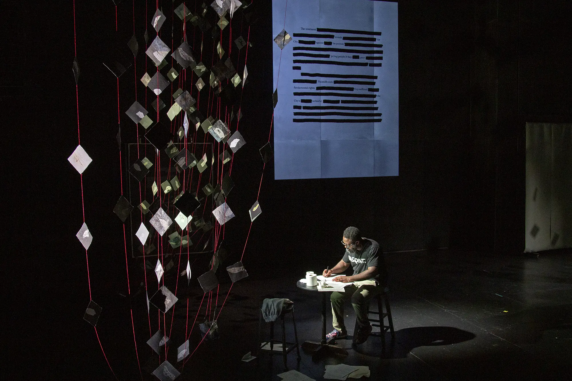 Freedom Reads CEO & Founder Reginald Dwayne Betts sits under the spotlight scribbling words on paper in a dramatic recreation of the moment he became a poet in a scene from his solo show FELON: An American Washi Tale.